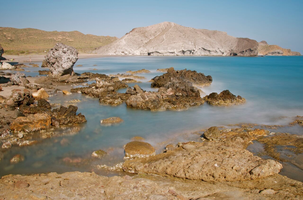 Rocas en la orilla de la Cala de la Media Luna, con el agua clara y los cerros detrás