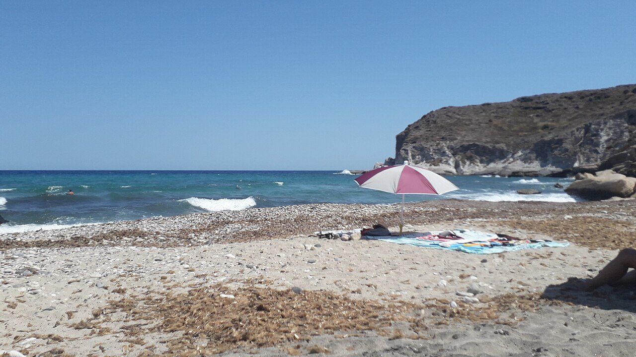 Orilla de piedras y arena de la Cala del Plomo, con el acantilado al fondo